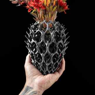 Intricate black 3D-printed pineapple vase holding colorful flowers on black background.