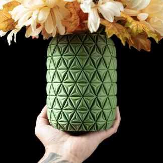 A detailed shot of a 3D-printed green vase with intricate geometric patterns, held by a tattooed hand against a black background.